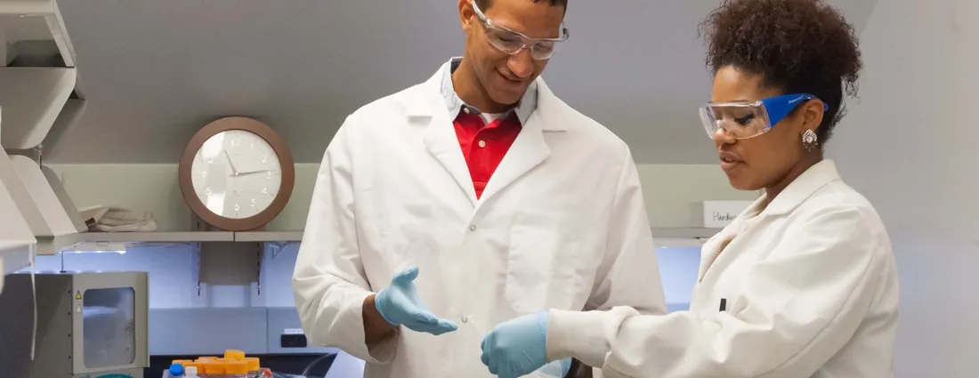 Two researchers wearing lab coats, safety glasses, and gloves collaborate on an experiment in a laboratory while examining a sample or research materials.