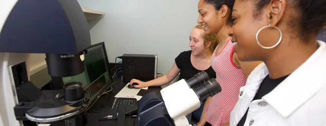 Three students work together in a research laboratory, viewing microscope imaging results on a computer while using advanced microscopy equipment.
