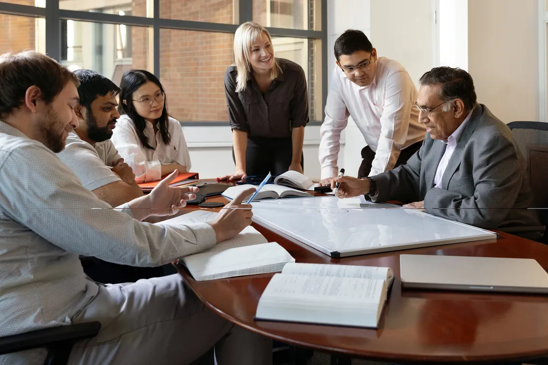 Individuals in business formal stand and sit around a conference table collaborating on a white board surrounded by paper printouts.
