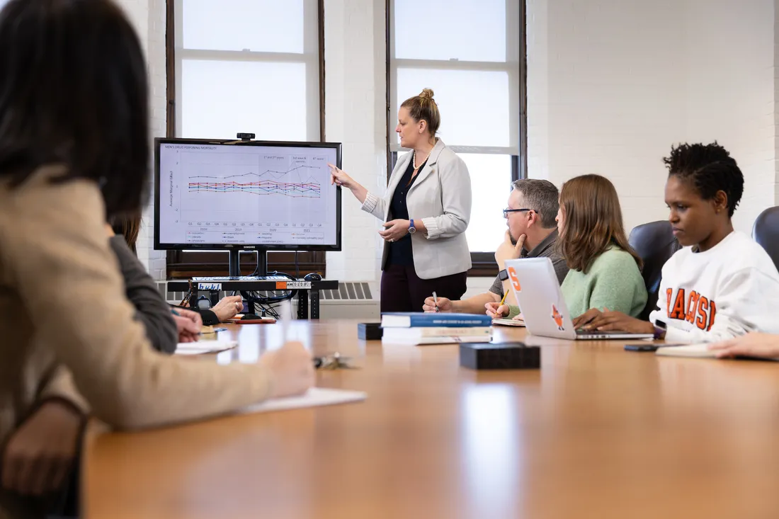 Woman stands at front of conference room pointing to data on large monitor as people sitting around large conference room table look on.