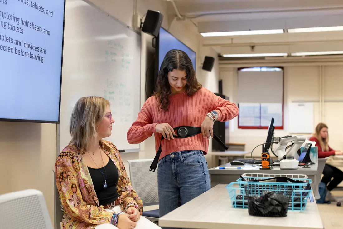 Two individuals interact in research lab. Sitting individual looks at standing individual who is holding a belt-like instrument against ribs.