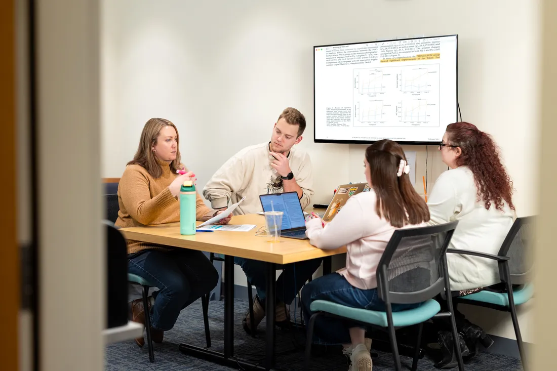 Four people sit at work table in office meting room. There is a laptop on the table and a tv monitor on the wall with information from the laptop projected on it.