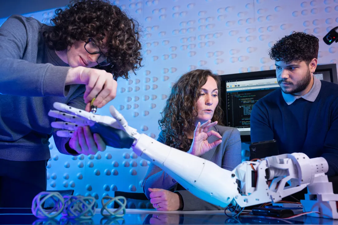 Two people look on and discuss as third person tools adjustment to bionic arm.