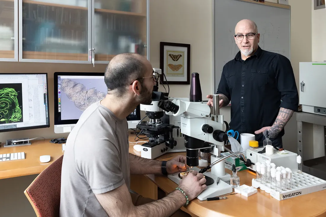 Two researchers work in a laboratory, with one seated at a workstation and the other standing beside lab equipment while discussing research activities.