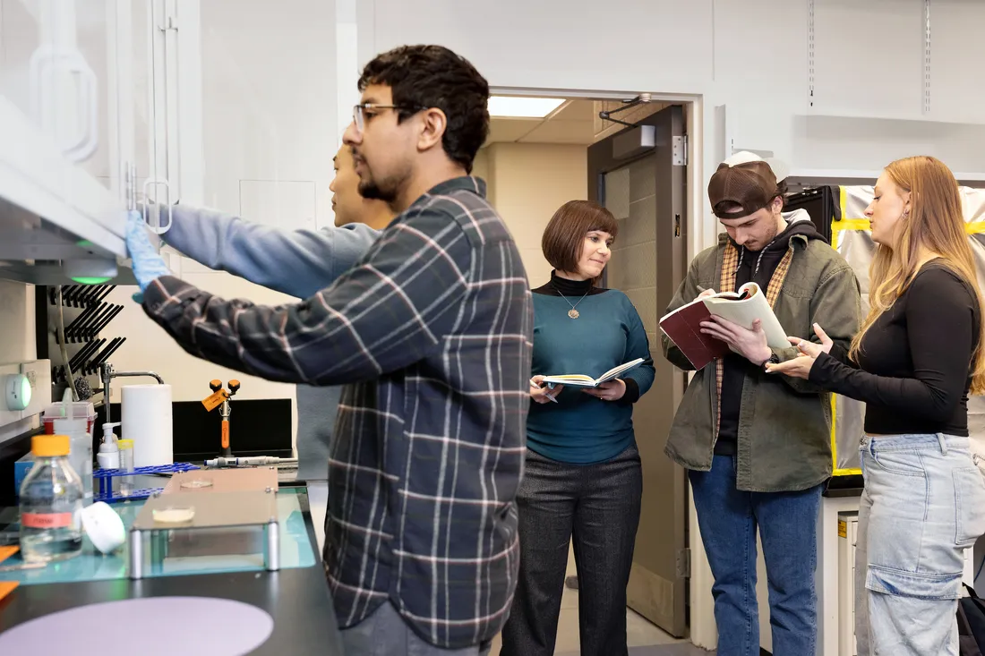 People in lab have conversation over books while person at counter points to lab equipment.