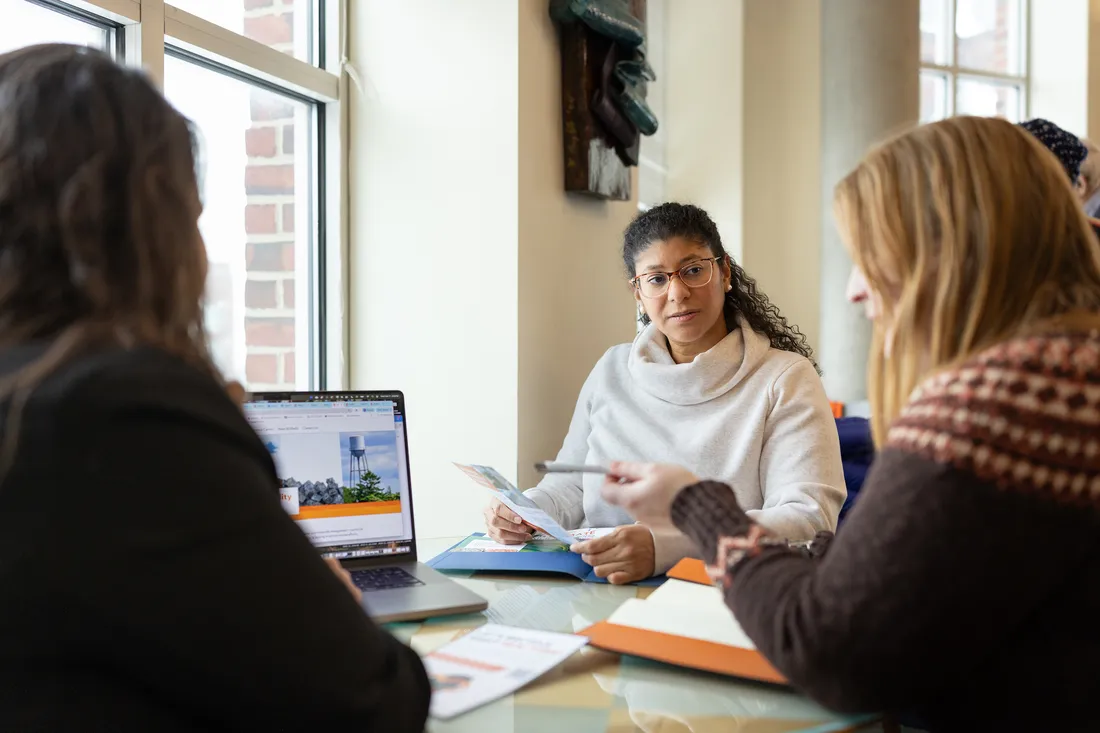 Three women sit together around table in open working space with papers and open laptop on table.