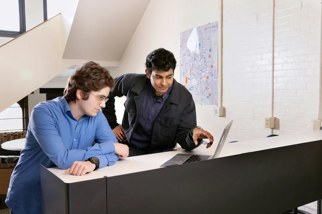 Two people working together at a desk, looking at information on a laptop in an office or research setting.